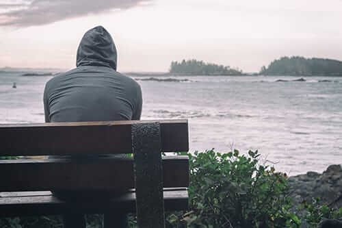 A person with their hood up sits on a bench overlooking the ocean to represent depression and anxiety