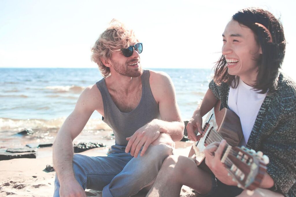 Two young men sitting on the beach, smiling and enjoying time together; one playing guitar, emphasizing friendship and support in recovery.