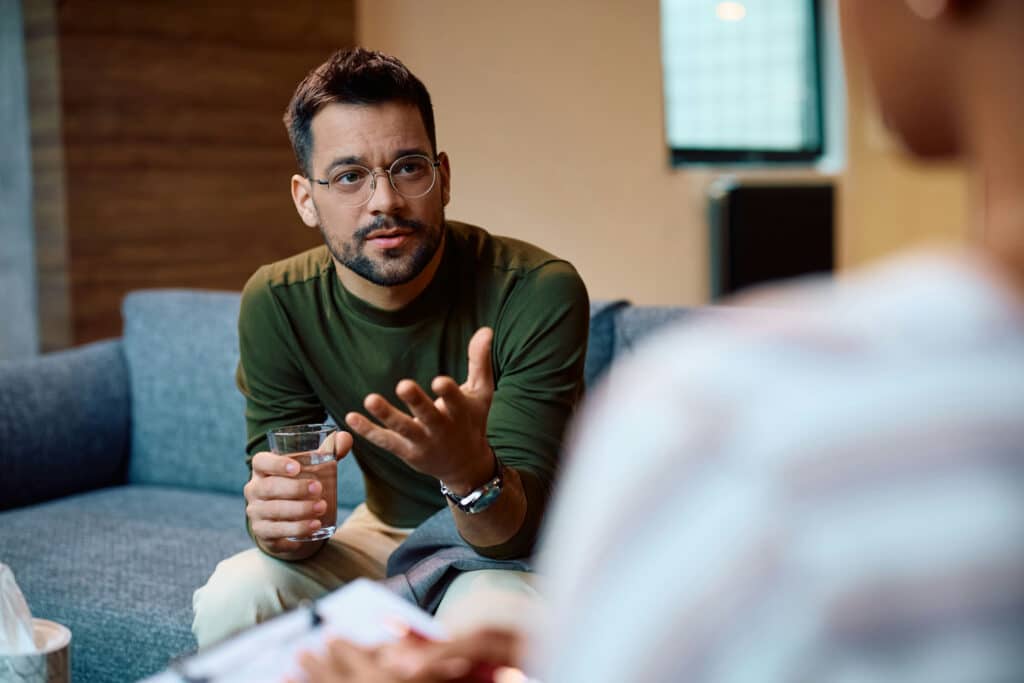 Man discussing benzodiazepine risks in therapy session, holding a glass of water, seated on a couch, with a notepad visible in foreground.