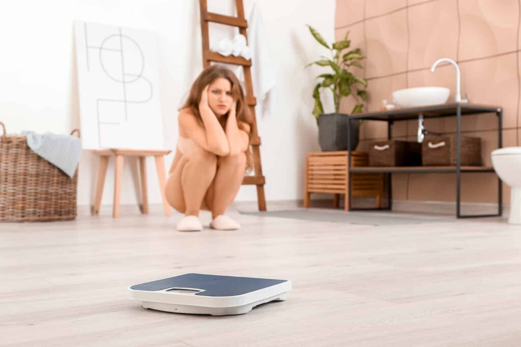 Young woman sitting on the floor, visibly distressed, with a weighing scale in the foreground, illustrating the mental struggles associated with atypical anorexia and body image issues.