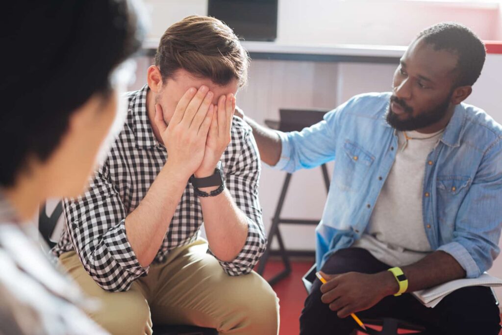 Group therapy session for addiction recovery, focusing on emotional support and connection, with a man in distress receiving comfort from a peer.