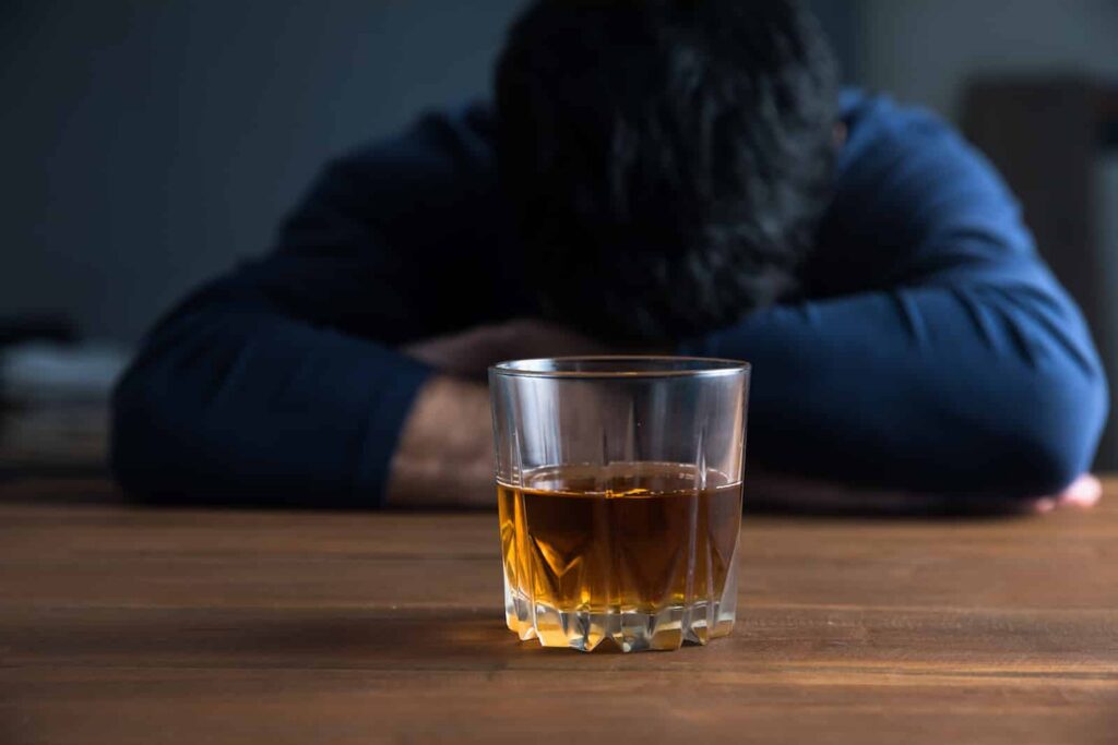 Man with head down on a table, glass of whiskey in front, symbolizing the dangers of mixing alcohol with Effexor for depression treatment.
