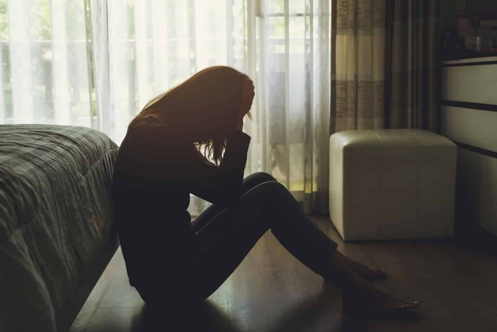 Woman sitting on the floor in a dimly lit room, holding her head in distress, reflecting the emotional struggles associated with heroin addiction and the challenges of recognizing substance use.