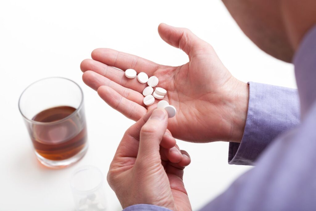 Person holding white pills in one hand, contemplating their use with a glass of brown alcohol nearby, illustrating the risks of mixing Xanax and alcohol.