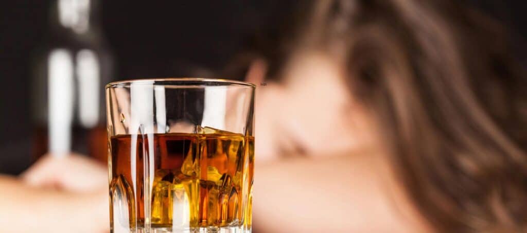 Glass of whiskey with ice in foreground, blurred figure of a person resting their head on a table in the background, symbolizing the effects of alcohol on health and blood thinning.