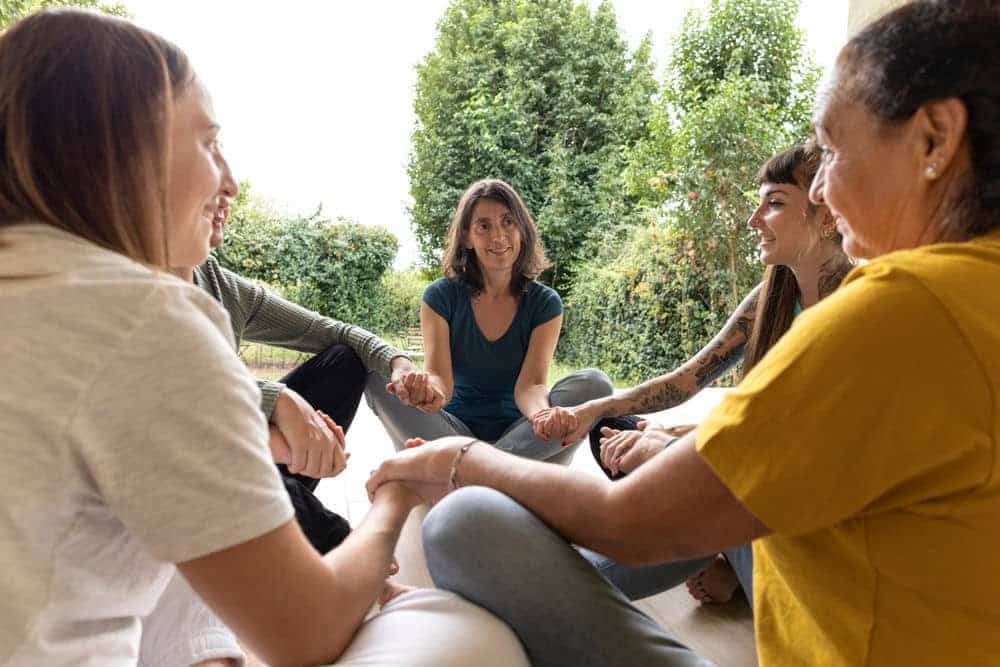 Small group of adults sitting in a circle holding hands during a supportive therapy or wellness session outdoors.