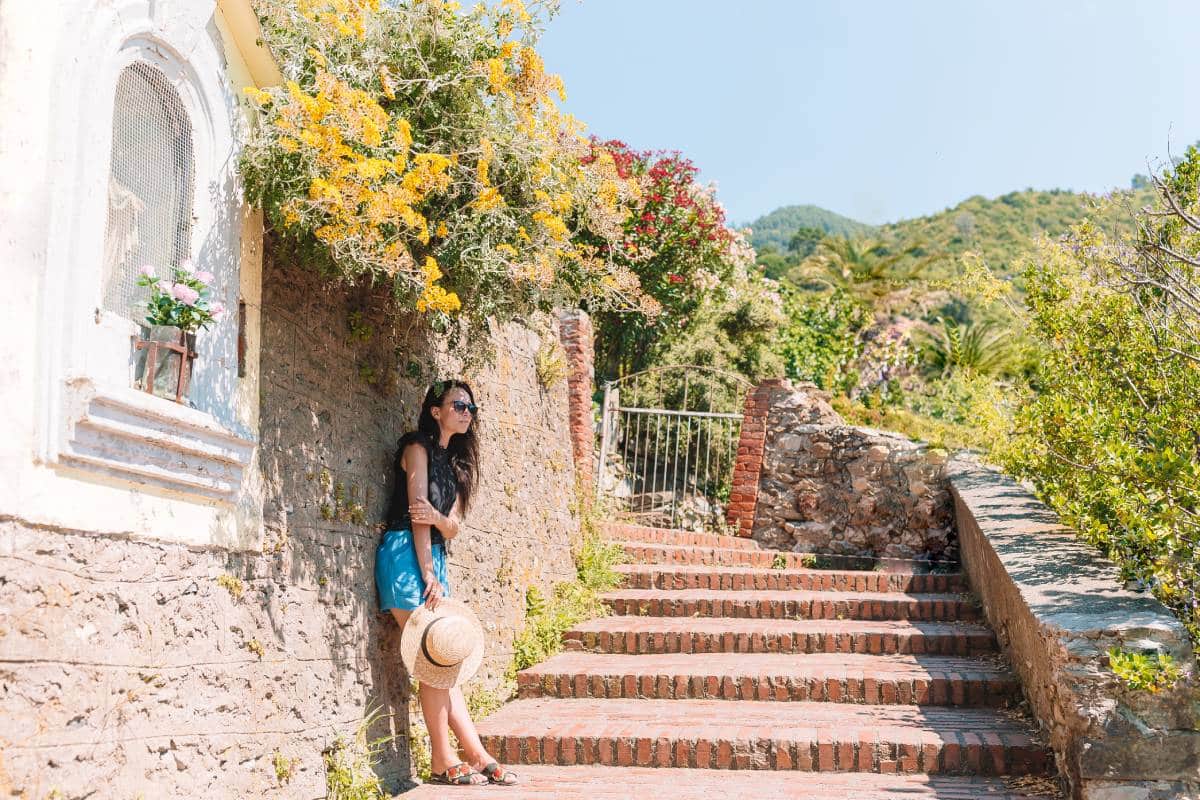 Woman leaning against a stone wall adorned with flowers, holding a straw hat, beside a brick pathway leading to a gated entrance, suggesting a serene outdoor setting in San Juan Capistrano.
